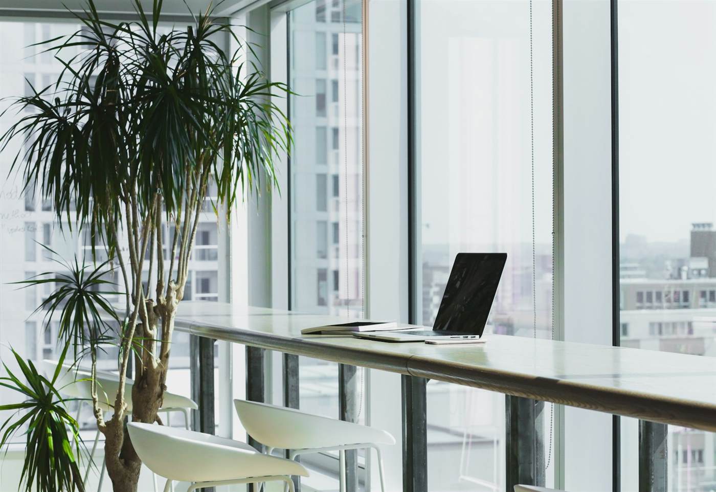A laptop and a closed notebook on a high table beside tall office windows