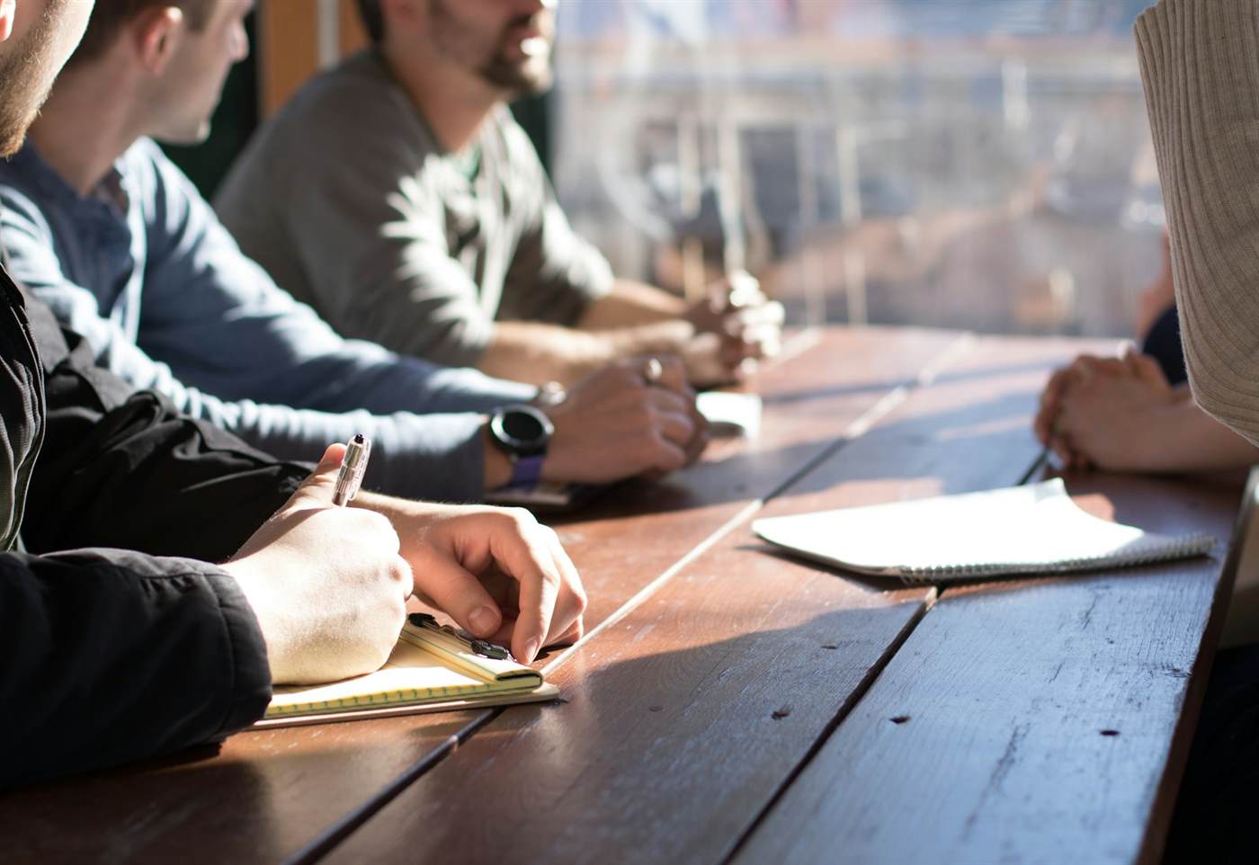 People seated along a wooden table, one of them writing in a notebook in daylight